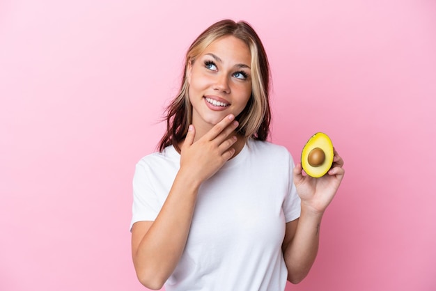 Jonge Russische vrouw met avocado geïsoleerd op een roze achtergrond terwijl ze glimlacht