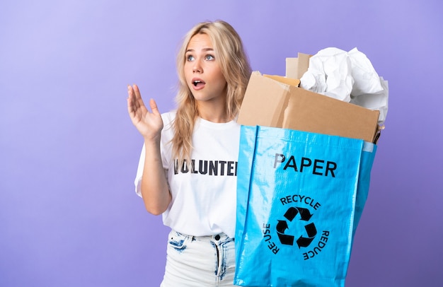 Jonge Russische vrouw met een recyclingzak vol papier om te recyclen geïsoleerd op paars met verrassende gelaatsuitdrukking