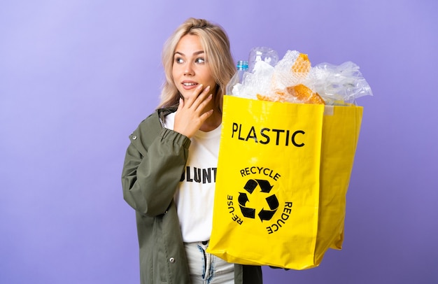 Jonge russische vrouw met een recyclingzak vol papier om te recyclen geïsoleerd op paars opzoeken tijdens het glimlachen