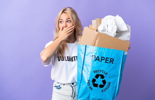 Jonge Russische vrouw met een recyclingzak vol papier om te recyclen geïsoleerd op paarse muur doet verrassingsgebaar terwijl ze naar de zijkant kijkt