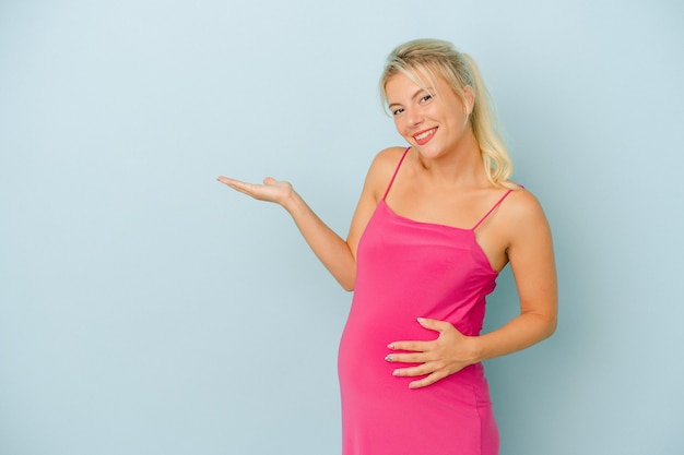 Jonge Russische vrouw zwanger geïsoleerd op blauwe achtergrond met een kopie ruimte op een palm en met een andere hand op de taille.