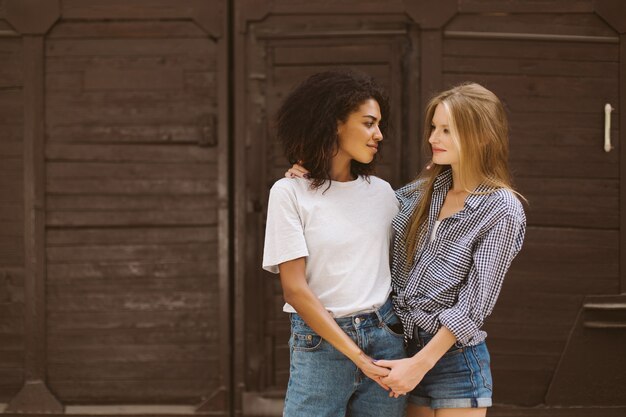 Jonge schattige Afro-Amerikaanse vrouw met donker krullend haar in T-shirt en spijkerbroek en vrouw met blond haar in shirt en denim shorts dromerig kijken elkaar tijd samen buiten doorbrengen