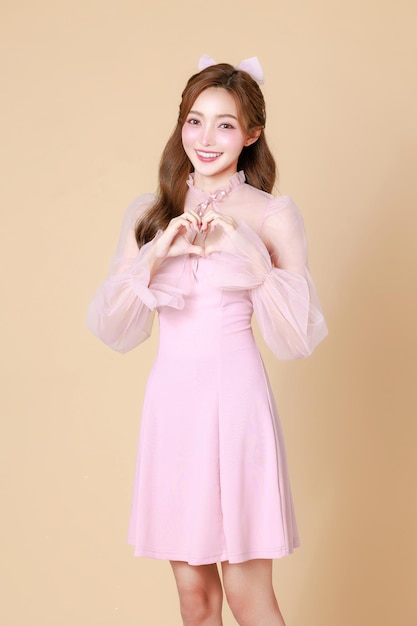 Jonge schattige Aziatische vrouw in een roze elegante jurk Koreaanse stijl make-up gehydrateerd gladde perfecte huid show liefde teken op een beige achtergrond gezichtsbehandeling cosmetologie plastische chirurgie