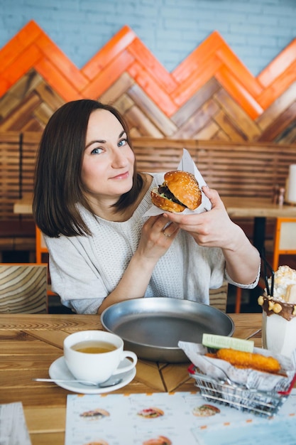 Jonge schattige brunette vrouw die een vers bereide heerlijke hamburger eet in het café. Tonen.