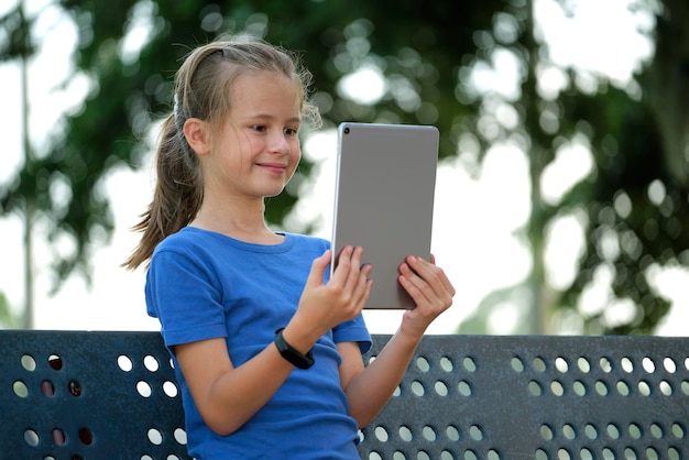 Jonge schattige lachende tienermeisje kijkend naar het scherm van een digitale tablet zittend op een bankje in het park op zonnige zomerdag