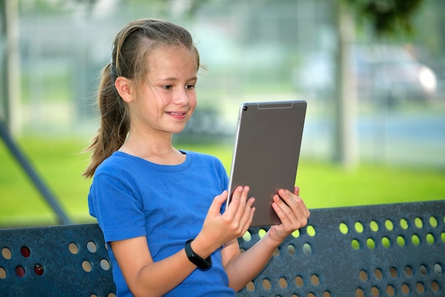 Jonge schattige lachende tienermeisje kijkend naar het scherm van een digitale tablet zittend op een bankje in het park op zonnige zomerdag