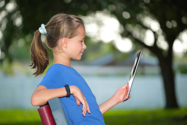 Jonge schattige lachende tienermeisje kijkend naar het scherm van een digitale tablet zittend op een bankje in het park op zonnige zomerdag