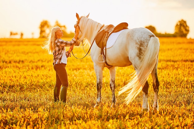 Jonge, schattige, vrolijke, tevreden glimlachende vrouw die een mooi wit paard omhelst en streelt op de weide bij zonsondergang op het gouden uur