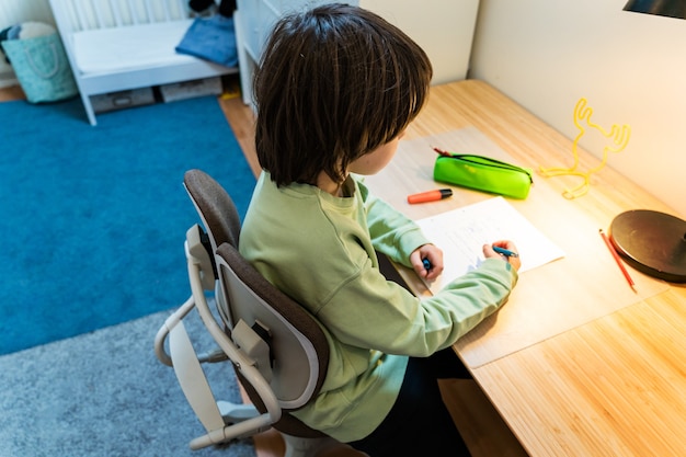 Jonge school jongen zijn huiswerk zittend op orthopedische stoel aan de tafel thuis. geconcentreerde schrijfoefeningen voor kinderen. thuisonderwijs concept.