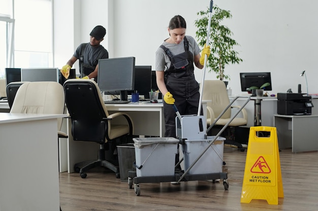 Jonge schoonmaakster die dweil in emmer knijpt terwijl ze de vloer op kantoor gaat wassen