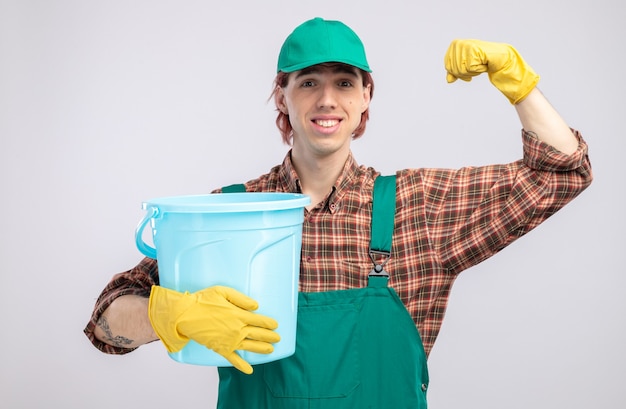 Jonge schoonmaakster in geruit hemd jumpsuit en pet met rubberen handschoenen die emmer vasthoudt glimlachend zelfverzekerd opheffende vuist als een winnaar die over een witte muur staat