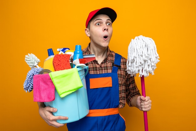 Jonge schoonmaakster met uniform en pet met emmer schoonmaakgereedschap met dweil geïsoleerd op oranje muur