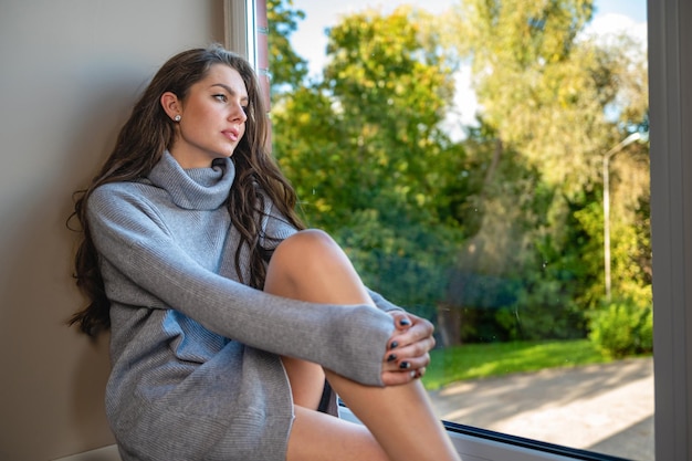 Jonge sensuele duizendjarige vrouw kijkt door het raam zittend op een vensterbank