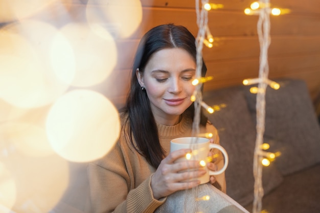 Jonge serene brunette vrouw met mok hete thee zittend op de bank bij een groot raam van het landhuis in het winterweekend en genietend van rust