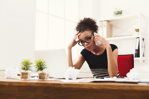 Jonge serieuze Afro-Amerikaanse zakenvrouw die telefonisch praat met papieren, zittend op een moderne kantoorwerkplek. Bedrijfsconsulting.