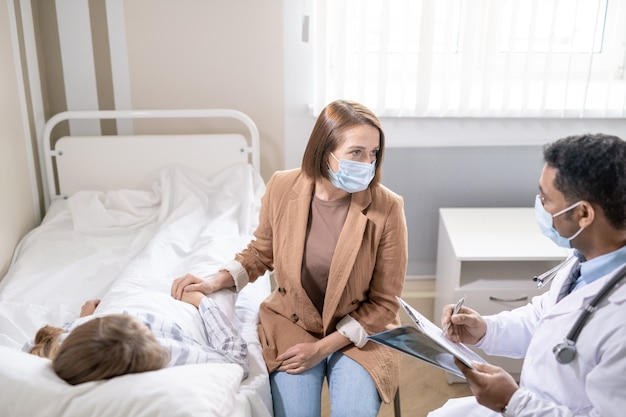 Jonge serieuze brunette vrouw met beschermend masker praat met dokter over haar zieke vriend die in bed ligt dichtbij in het hedendaagse covid-ziekenhuis