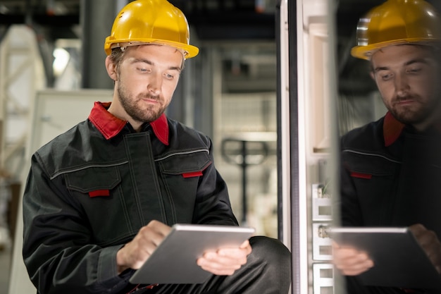 Jonge serieuze ingenieur in werkkleding en veiligheidshelm die door technische gegevens in digitale tablet bladert door een van de industriële machines