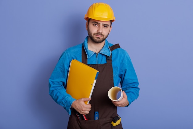 Jonge serieuze ingenieur met papieren mappen in handen, camera kijken met zelfverzekerde gezichtsuitdrukking, helm en schort dragen