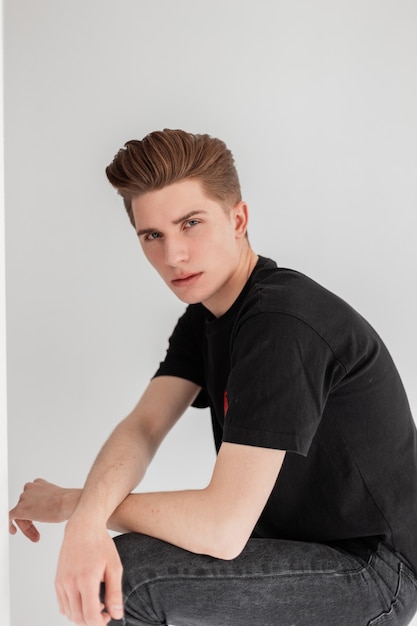 Jonge serieuze modieuze man met stijlvol kapsel in trendy casual zwart t-shirt voor jongeren uit de nieuwe zomercollectie staat in de buurt van vintage muur in studio. Knappe vogue man in vintage slijtage binnenshuis.