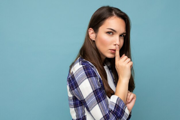 Jonge serieuze mooie brunet vrouw met oprechte emoties, gekleed in een trendy geruit hemd, geïsoleerd op een blauwe achtergrond met lege ruimte en een shhh-gebaar tonend. Houd geheim concept.