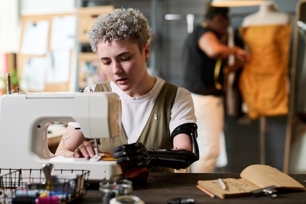 Foto jonge serieuze naaister met gedeeltelijke arm naaiende kleding op elektrische machine