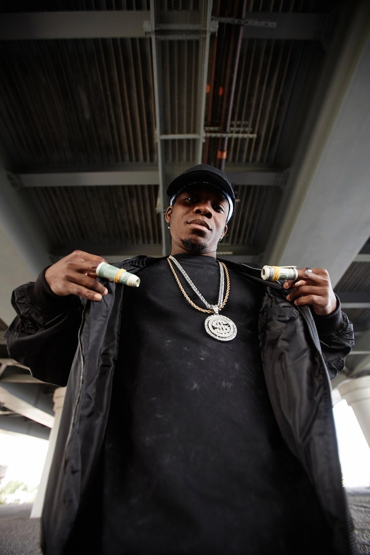 Foto jonge serieuze rapper met gerolde dollarbiljetten op zijn vingers staat voor de camera onder een brug in een stedelijke omgeving tijdens een optreden