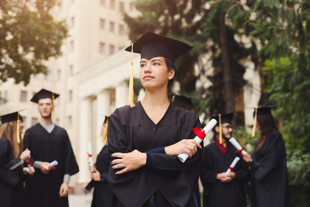 Jonge serieuze vrouw op haar afstudeerdag op de universiteit, staande met een multi-etnische groep studenten. Onderwijs, kwalificatie en toga concept.