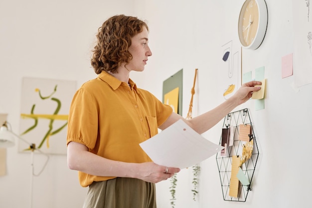 Jonge serieuze zakenvrouw die briefpapier met werktaken op de muur zet