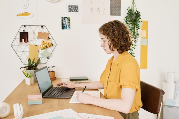 Jonge serieuze zakenvrouw in vrijetijdskleding die bij het bureau voor de laptop zit