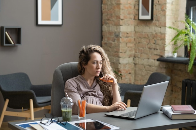 Jonge serieuze zwangere vrouw die verse wortel eet tijdens het werk