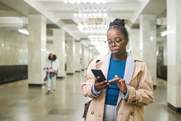 Jonge serieuze zwarte vrouw in trenchcoat die door locatiekaart in smartphone kijkt