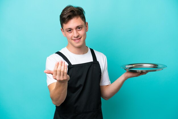 Jonge serveerster met dienblad geïsoleerd op een blauwe achtergrond die uitnodigt om met de hand te komen. Blij dat je gekomen bent