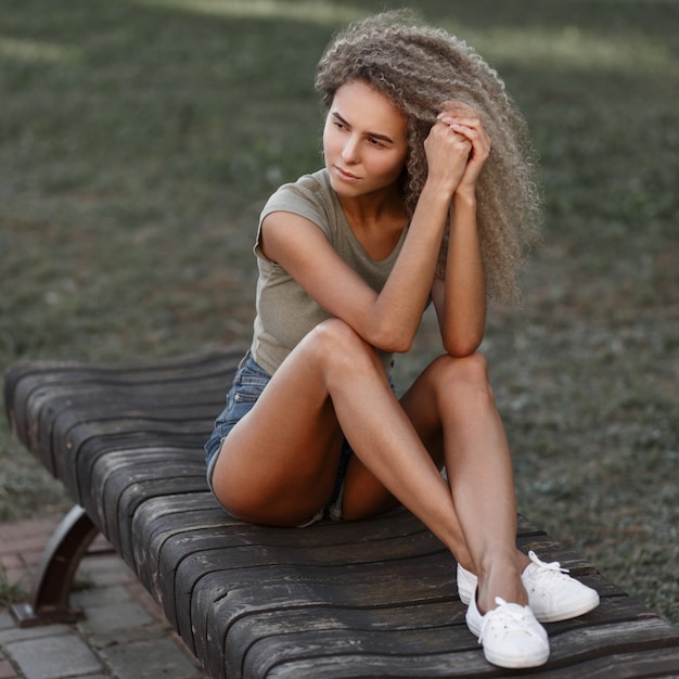 Jonge sexy mooie gekrulde vrouw in stijlvolle casual jeans kleding met korte broek en witte sneakers zit op een bankje in het park