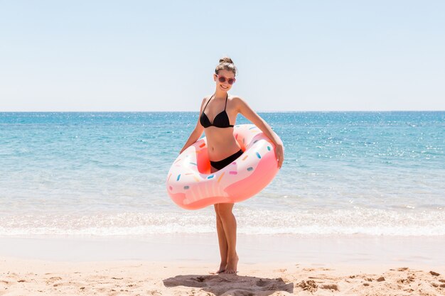 Jonge sexy mooie vrouw blijft op het zeestrand en speelt met opblaasbare ringdoughnut