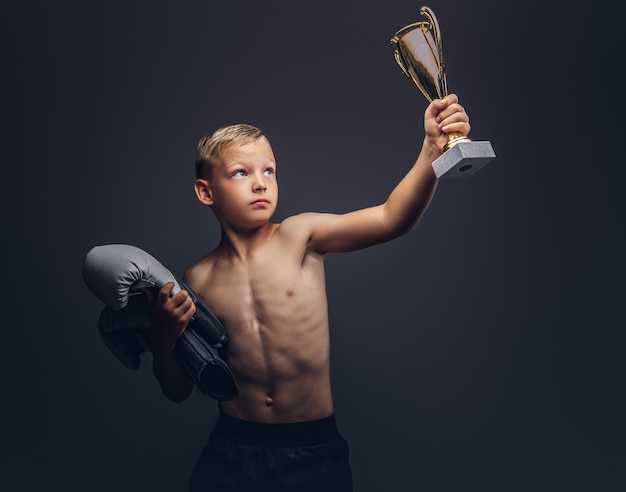 Jonge shirtloze jongen houdt bokshandschoenen en de winnaarsbeker vast. Geïsoleerd op een donkere achtergrond.