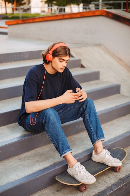 Jonge skater in oranje koptelefoon die bedachtzaam mobiele telefoon gebruikt die op trappen zit met klassiek skateboard in skatepark