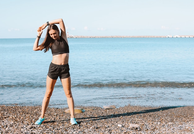 Jonge slanke atletische vrouw in sportkleding die oefeningen op het overzeese strand in ochtend doen