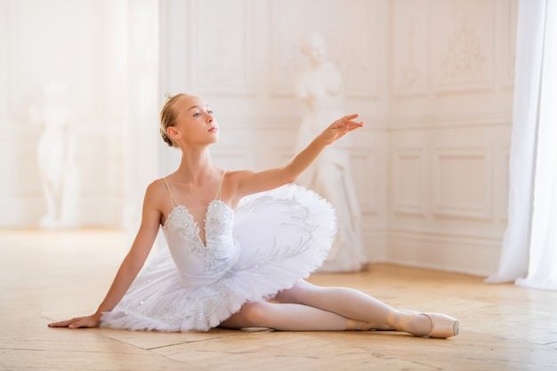 Jonge slanke ballerina in een witte tutu in pointe-schoenen zit in sierlijke pose op de vloer in grote mooie witte hal.