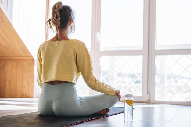 Jonge slanke fitness blonde vrouw oefent ochtendyoga bij het raam van huis