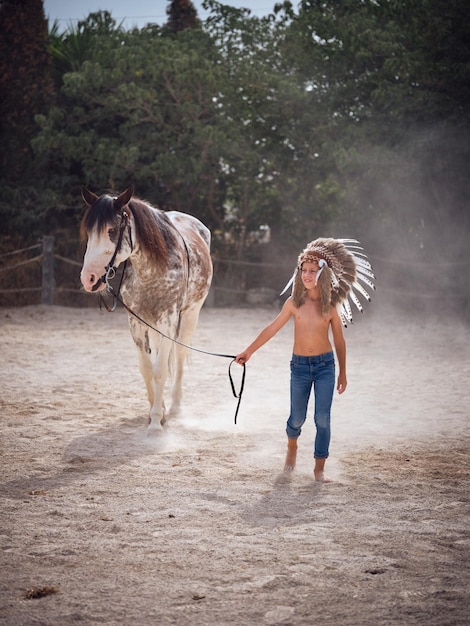 Jonge slanke jongen met hoofddeksel die de teugels van een paard vasthoudt terwijl hij op zandgrond tegen groene bomen slentert en wegkijkt