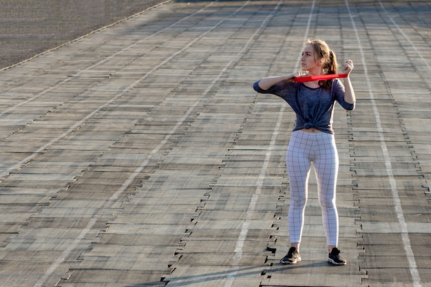 Jonge slanke vrouw in sportkleding die hurkzitoefening met elastiekje doen op een zwart met een laag bedekt stadionspoor