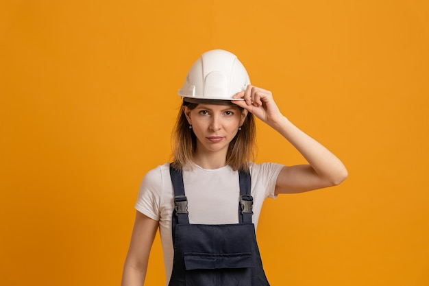 Jonge slanke vrouwenbouwer in een helm op een oranje achtergrond houdt met haar hand een harde hoed vast