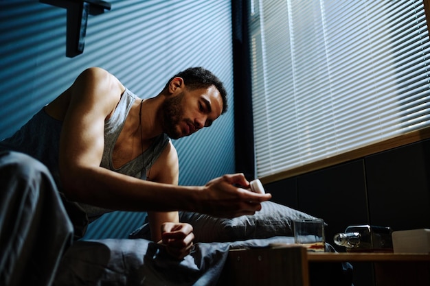 Jonge slapeloze man zittend op bed en kijkend naar fles pillen in zijn hand