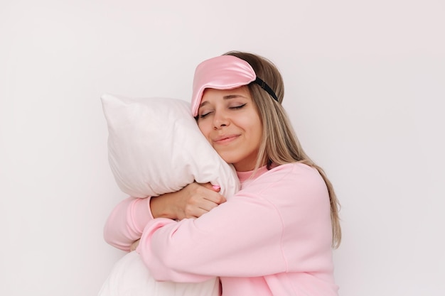 Jonge slaperige glimlachende blonde vrouw in roze pyjama en slaapmasker met gesloten ogen omhelst kussen