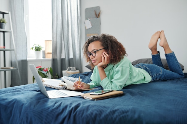 Jonge slimme student van gemengd ras in spijkerbroek en hoodie liggend op bed voor laptop en kijken naar online les terwijl hij aantekeningen maakt in schrift