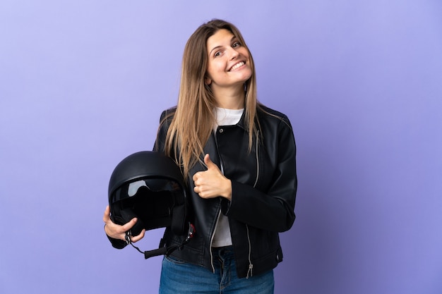 Jonge slowaakse vrouw die een motorhelm houdt die op purpere muur wordt geïsoleerd die een duim omhoog gebaar geeft