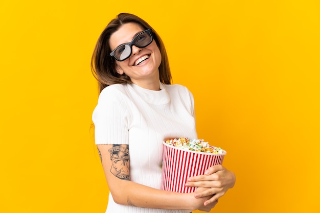 Jonge Slowaakse vrouw geïsoleerd op gele achtergrond met 3D-bril en met een grote emmer popcorn