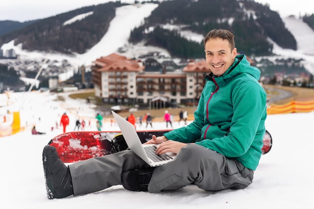 Jonge snowboarder met behulp van laptop op besneeuwde berg. Witte sneeuw en blauwe lucht. Kopieer ruimte voor advertentie.