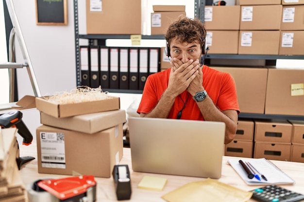 Jonge spaanse callcenter agent man aan het werk in magazijn geschokt mond bedekkend met handen voor fout geheim concept