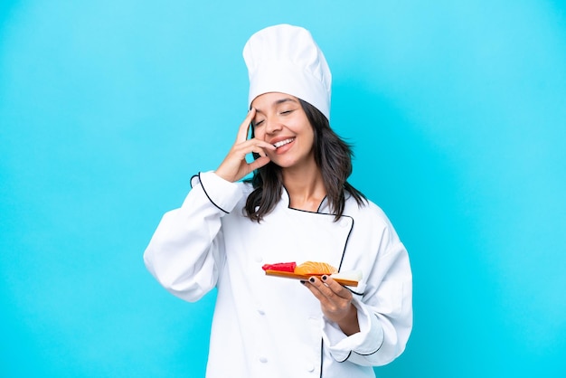 Jonge spaanse chef-kokvrouw die sashimi houdt die op blauwe achtergrond wordt geïsoleerd die veel glimlacht